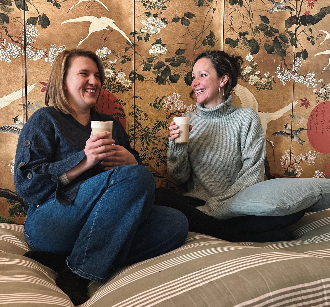 Britta und Kaltrina, die Gastgeberinnen von Our Shift, sitzen lachend mit Tee auf Bodenkissen vor einem goldenen Paravent